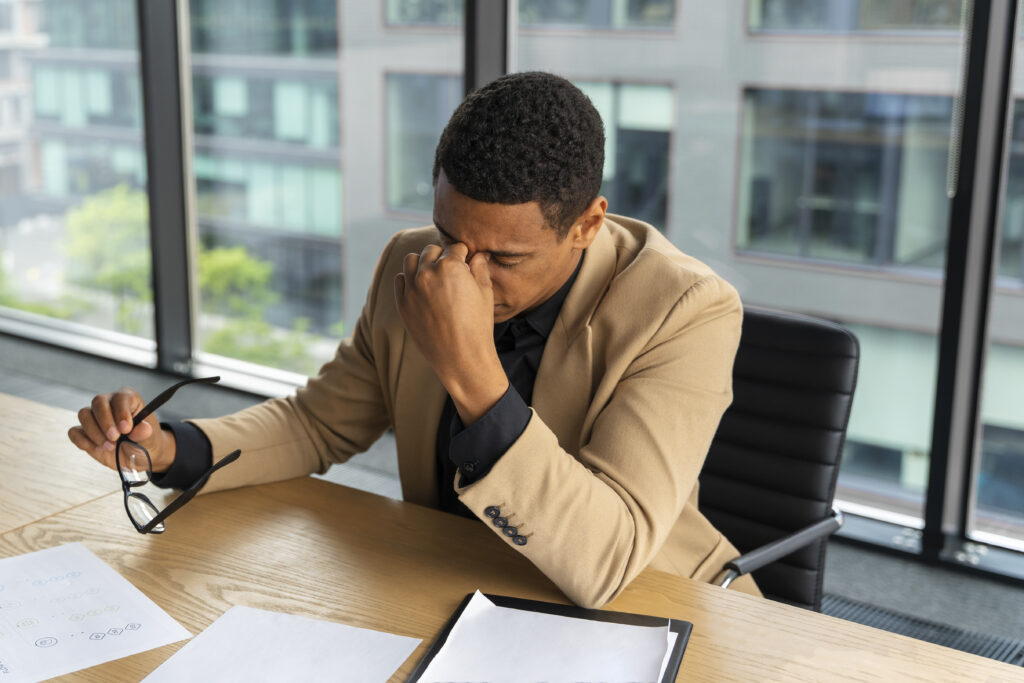 A man in a tan blazer sits at a desk with his head in his hand, looking stressed or tired. He is holding glasses in his other hand, and there are papers on the desk in front of him. The background shows a modern office building with large windows.
